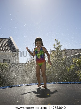 Full length of a little girl jumping on trampoline against water spray