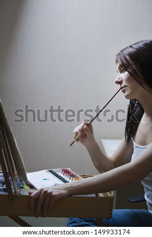 Side view of a woman sitting in front of easel and holding paintbrush