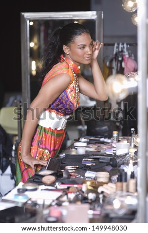 Side view of a young fashion model applying makeup in dressing room mirror
