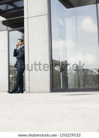 Full length of businessman using mobile phone while leaning on office wall