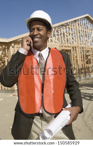 Happy African American worker with blueprints using cell phone at construction site