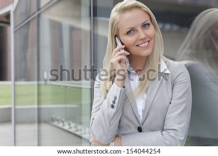 Portrait of young businesswoman communicating on cell phone while leaning on glass wall