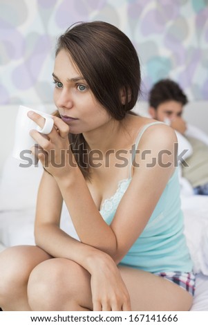 Young woman having coffee on bed with man lying in background