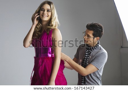 Happy model using cell phone while male designer adjusting her dress in studio