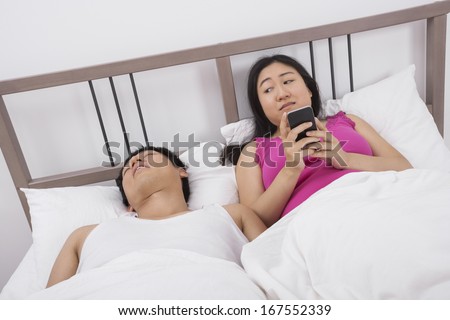 Woman using cell phone while looking at man sleeping in bed