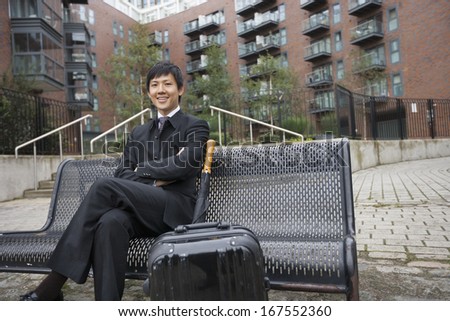 Portrait of confident businessman with luggage sitting on bench against buildings