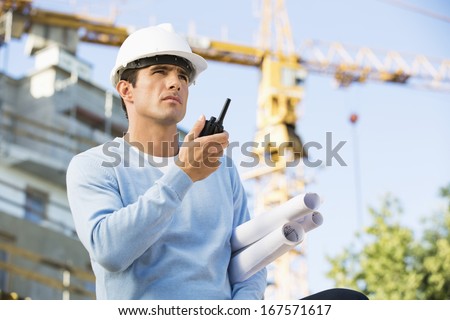 Male architect with blueprints using walkie-talkie at construction site