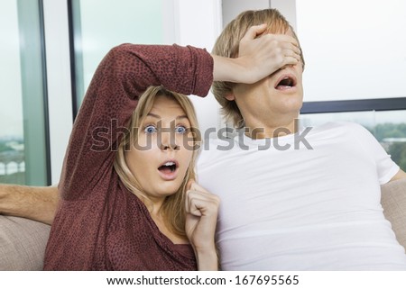 Shocked woman covering man's eyes while watching TV at home