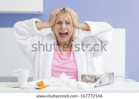 Portrait of young exhausted woman screaming while suffering from headache and cold in kitchen