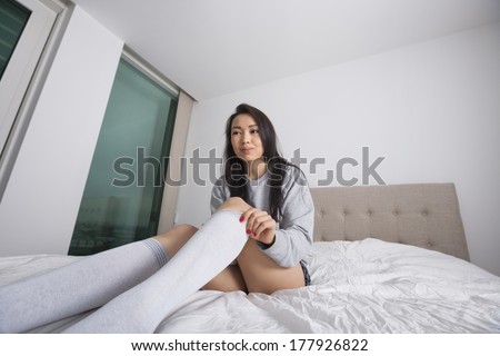 Thoughtful young woman putting on knee socks on bed