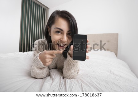Portrait of happy woman pointing at smart phone while lying in bed