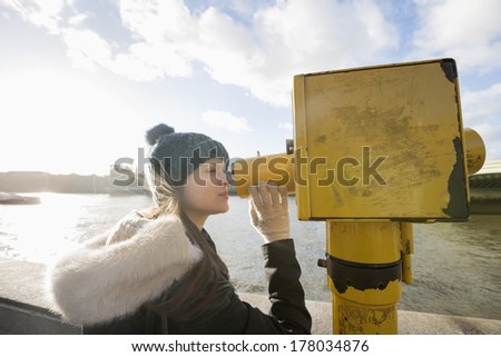 Side view of young woman in hooded sweatshirt looking through telescope by river Thames; London; UK
