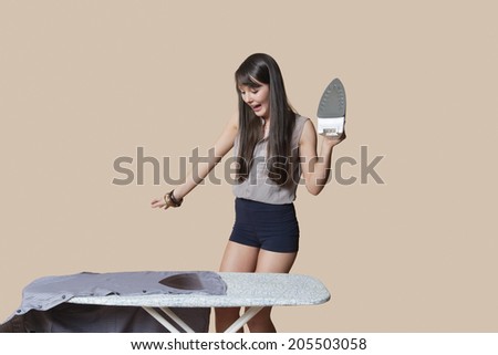 Shocked young woman looking at burnt shirt on ironing board over colored background