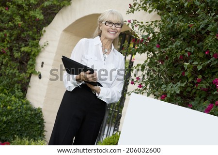 Cheerful senior woman standing by sign board with clipboard while looking away