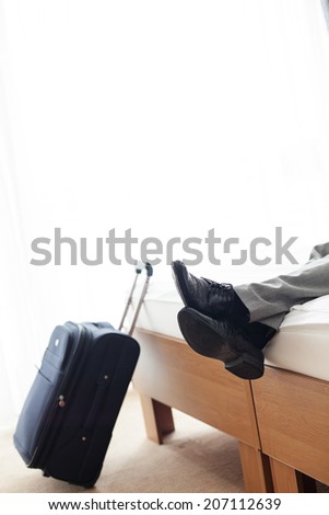 Low section of businessman lying on bed beside luggage in hotel room