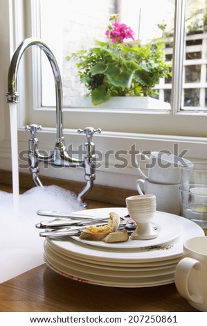 Closeup of a stack of dirty dishes and silverware by sink with running water below window with flower