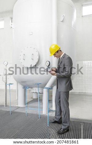 Full length side view of young male supervisor with clipboard standing by storage tank in industry