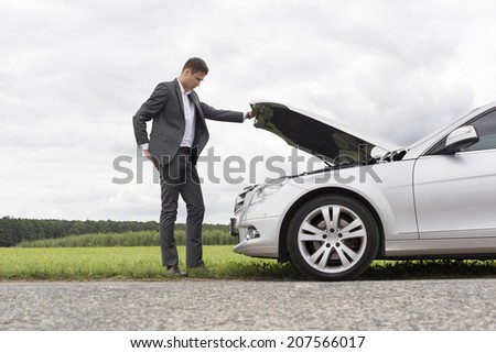 Full length side view of young businessman examining broken down car engine at countryside