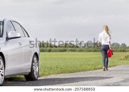 Full length rear view of woman carrying gas can leaving behind broken down car at countryside