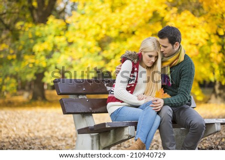 Passionate young man hugging shy woman on park bench during autumn