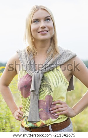 Thoughtful young woman looking away while standing with hands on hips in field