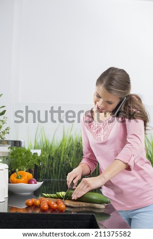 Smiling woman using cell phone while cutting cucumber at kitchen counter