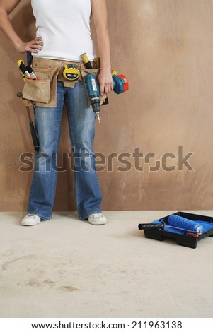 Lowsection of a young woman with toolbelt and drill leaning against wall