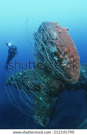 Underwater shoot of female scuba diver swimming near propeller of sunken battleship
