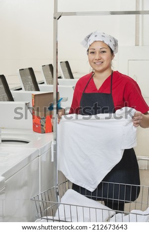 Portrait of a happy woman wearing uniform holding clothes with washing machines in background