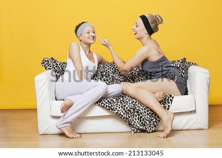 Happy young woman applying face pack on friend\'s face while sitting on sofa against yellow wall
