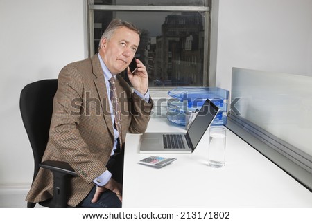 Middle-aged businessman using cell phone in front of laptop at desk in office