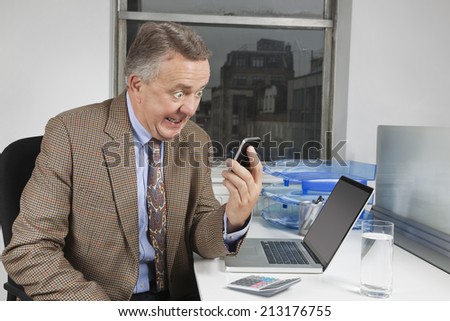 Angry middle-aged businessman looking at cell phone in office
