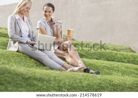 Full length of happy businesswomen looking at laptop while sitting on grass steps against wall