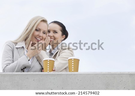 Happy businesswomen with disposable coffee cups sharing secrets against clear sky