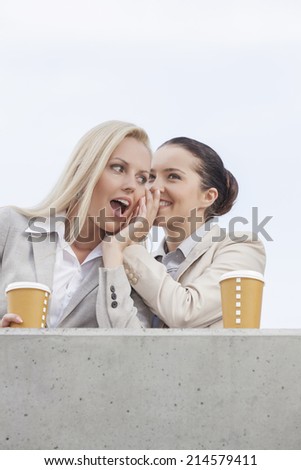 Low angle view of happy businesswomen with disposable coffee cups sharing secrets while standing on terrace against sky