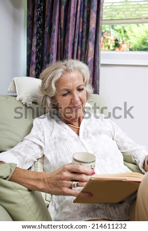 Senior woman with coffee cup reading book while relaxing on armchair at home