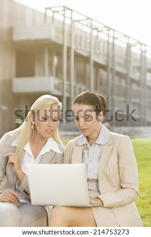 Young businesswomen using laptop together while sitting against office building