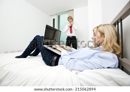 Businesswoman using laptop in bed with businessman adjusting tie at hotel
