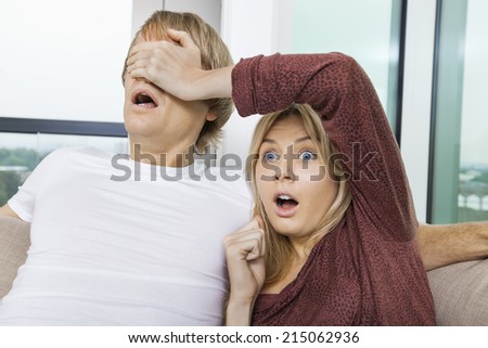Shocked woman covering man\'s eyes while watching TV at home