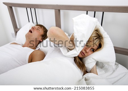 Frustrated woman covering ears with pillow while man snoring in bed