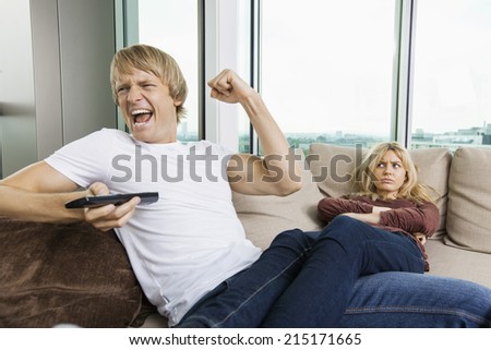 Angry woman staring at cheerful man as he watches TV in living room at home