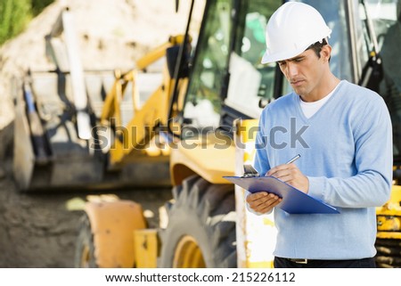 Male architect writing on clipboard against earthmover at construction site