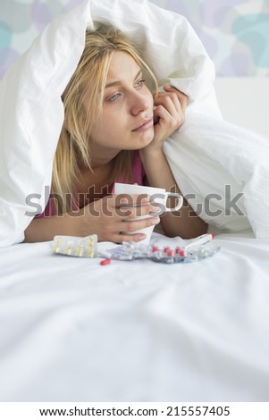 Young woman with coffee mug and medicines suffering from fever while covered in quilt on bed