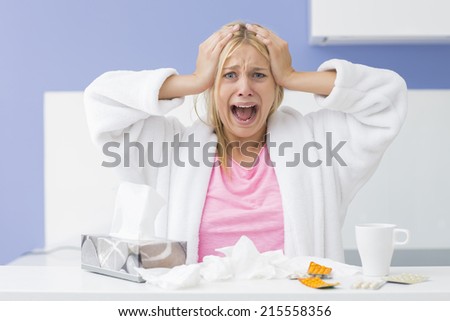 Portrait of young exhausted woman screaming while suffering from headache and cold in kitchen