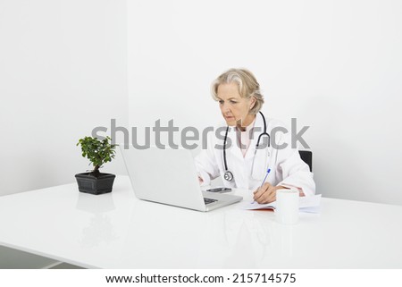 Senior female doctor writing notes while looking at laptop in clinic