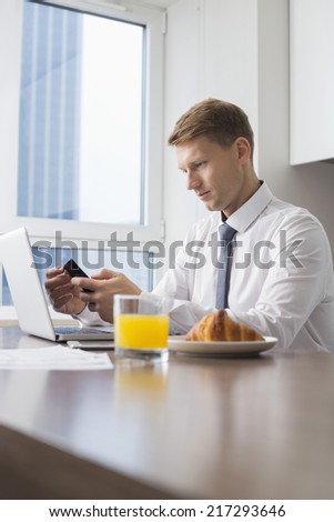 Mid adult businessman using cell phone with laptop on breakfast table