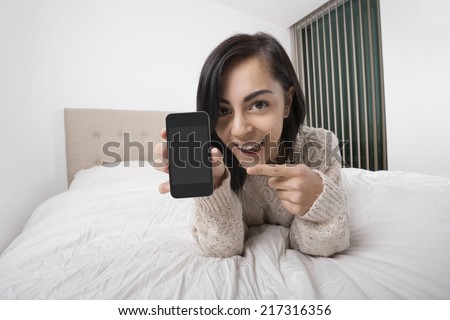 Portrait of happy woman pointing at smart phone while lying in bed