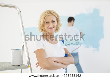 Portrait of woman holding paint roller with man painting wall in background