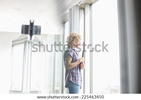 Side view of creative businesswoman holding files while looking through window in office