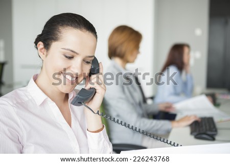 Smiling young businesswoman using landline telephone with colleagues in background at office